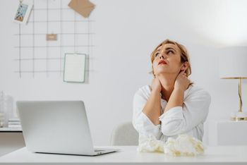 Free A stressed woman in white long sleeves sits at a desk with a laptop, looking upwards in an office. Stock Photo